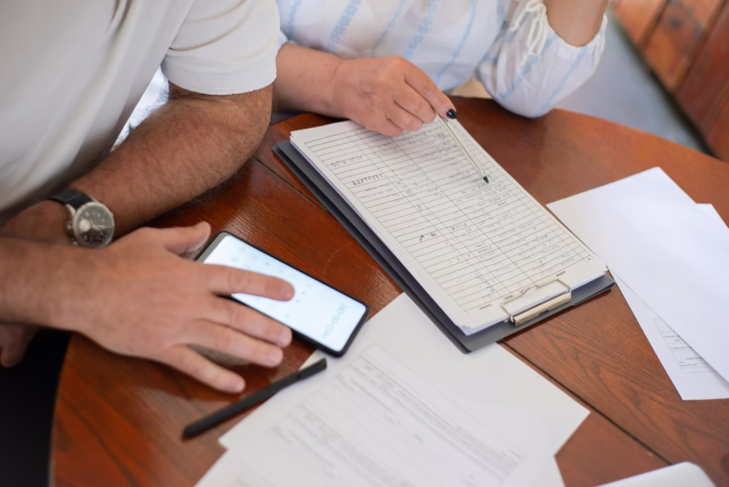 Senior couple examining bills and documents with smartphone and clipboard at home.